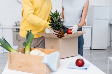 Cropped view of couple in latex gloves holding fresh fruits near boxes and rags in kitchen 