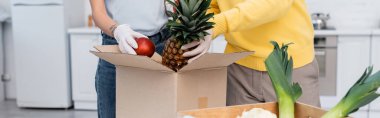 Cropped view of couple in latex gloves taking fruits from box in kitchen, banner 