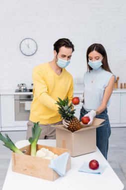 Young couple in latex gloves and medical masks taking fruits from carton box near rags in kitchen 