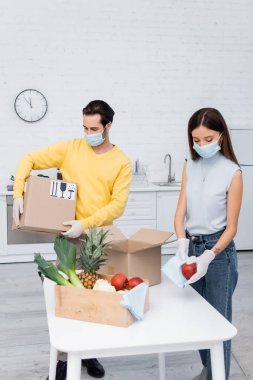Woman in medical mask cleaning apple near fruits and boyfriend holding carton box in kitchen 