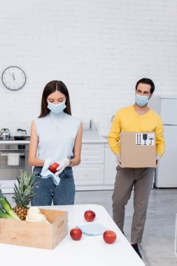 Man in medical mask holding carton box with signs near girlfriend cleaning apple and food in kitchen 