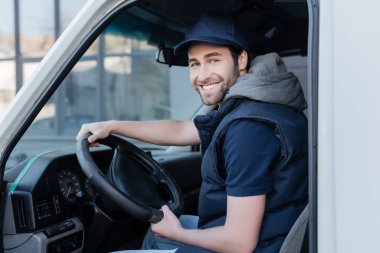 Smiling delivery man looking at camera near steering wheel in car 