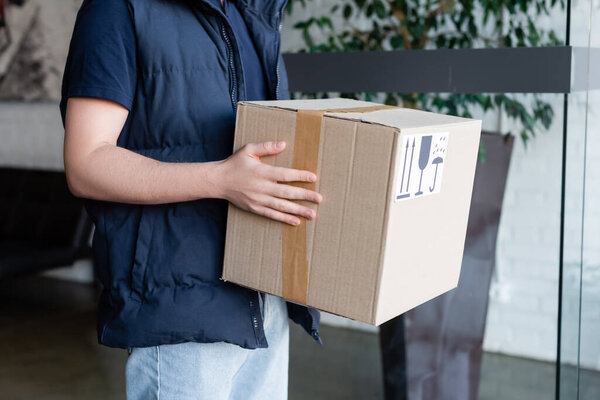 Cropped view of delivery man holding cardboard box with symbols in hallway 