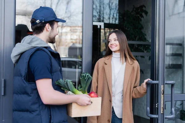 Smiling woman looking at courier with fresh food near door of building outdoors 