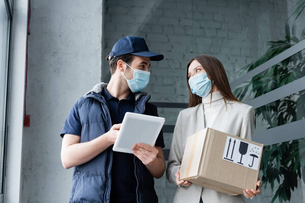 Delivery man in medical mask holding digital tablet near woman with box in hallway 