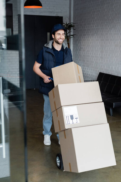 Positive courier standing near cart with packages in hallway 