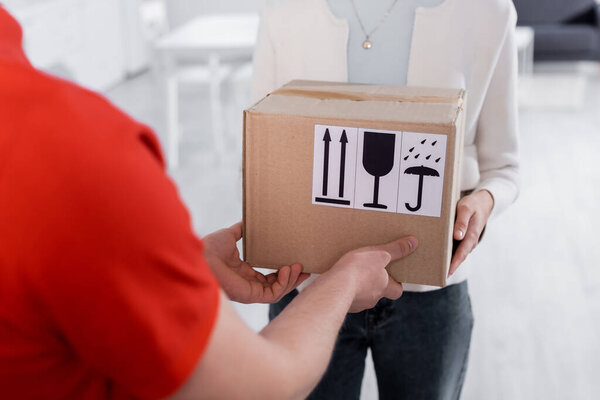 Cropped view of courier giving carton box with symbols to woman in hallway 
