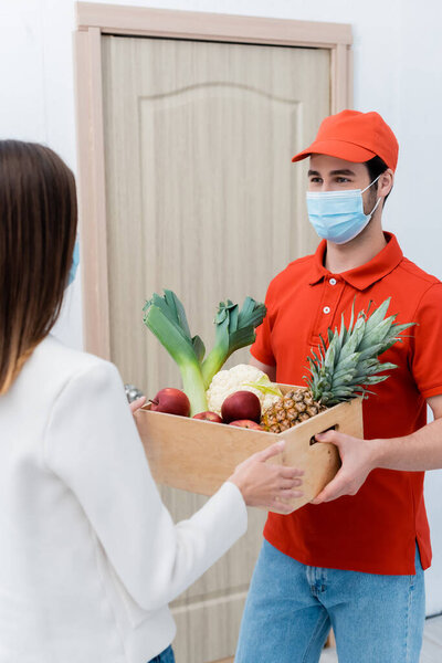 Delivery man in protective mask holding wooden box with fresh food near customer in hallway 