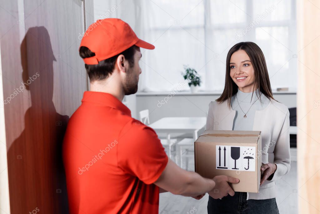 Blurred courier giving carton box to smiling woman in hallway 