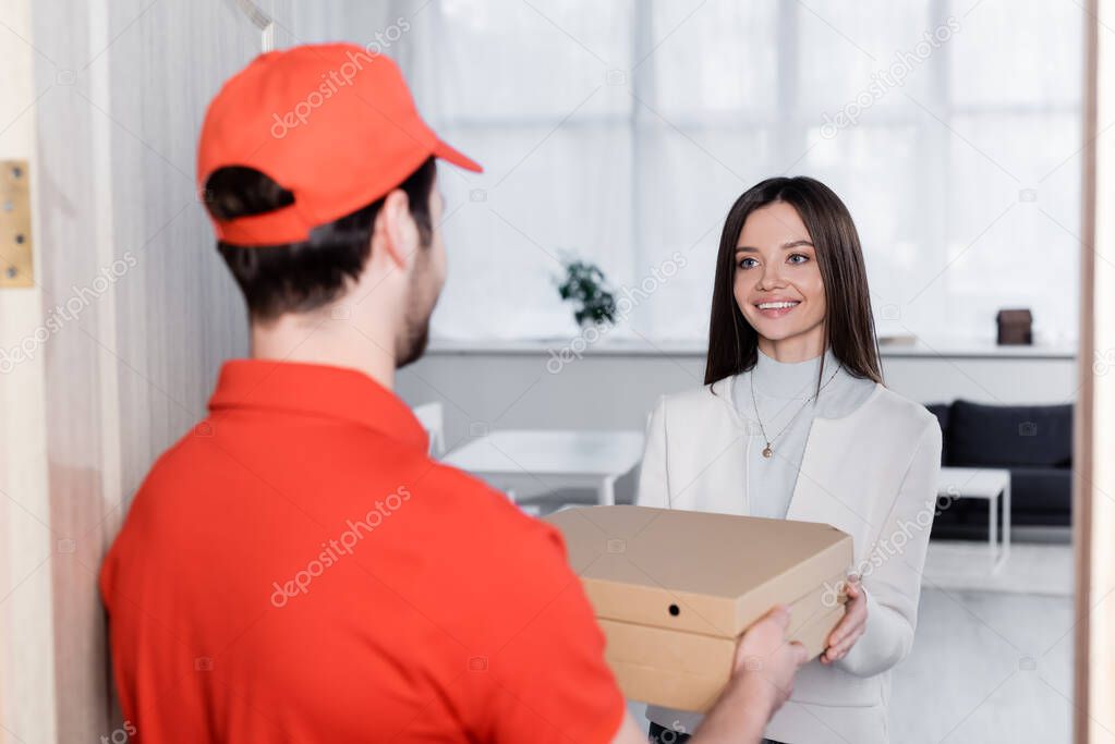 Positive woman taking pizza boxes from blurred courier in hallway 