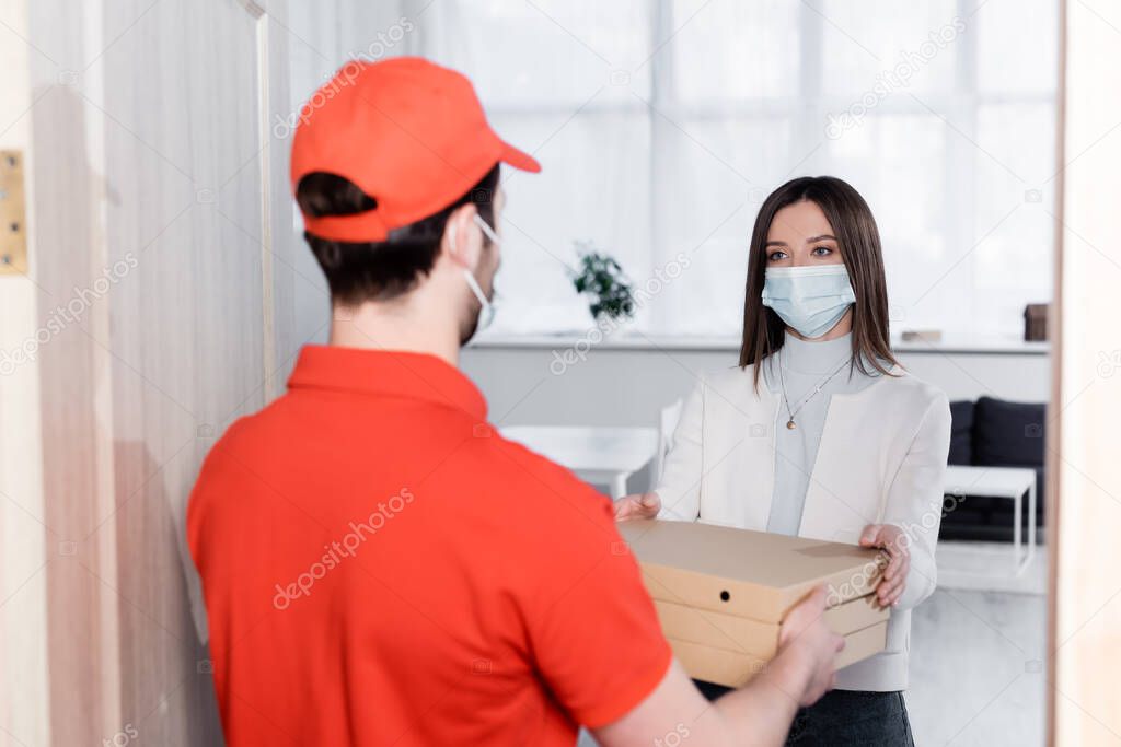 Woman in medical mask taking pizza boxes from blurred delivery man in hallway 