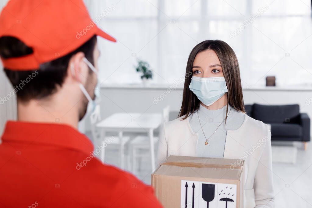 Young woman in medical mask holding carton box near blurred delivery man in hallway 