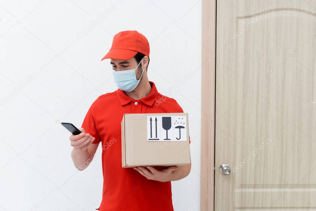 Delivery man in medical mask using smartphone and holding carton box in hallway 