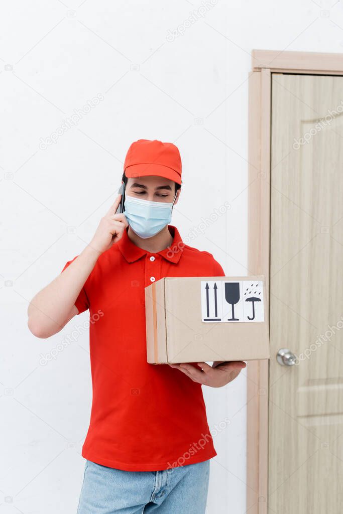 Courier in medical mask talking on smartphone and holding cardboard box in hallway 