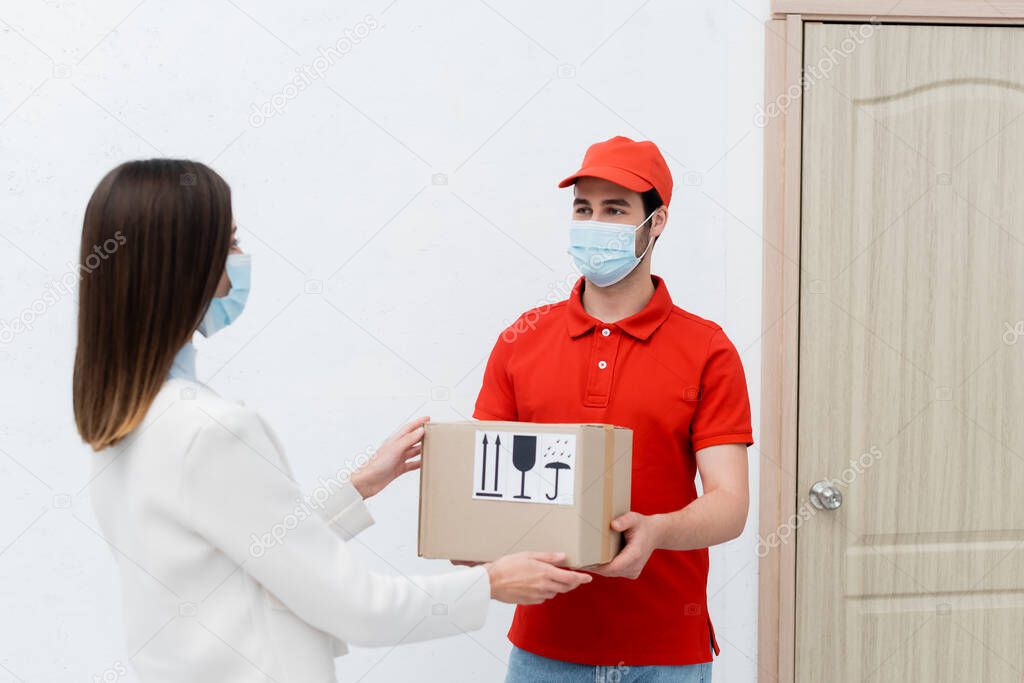 Delivery man in medical mask holding carton box near woman and door in hallway 