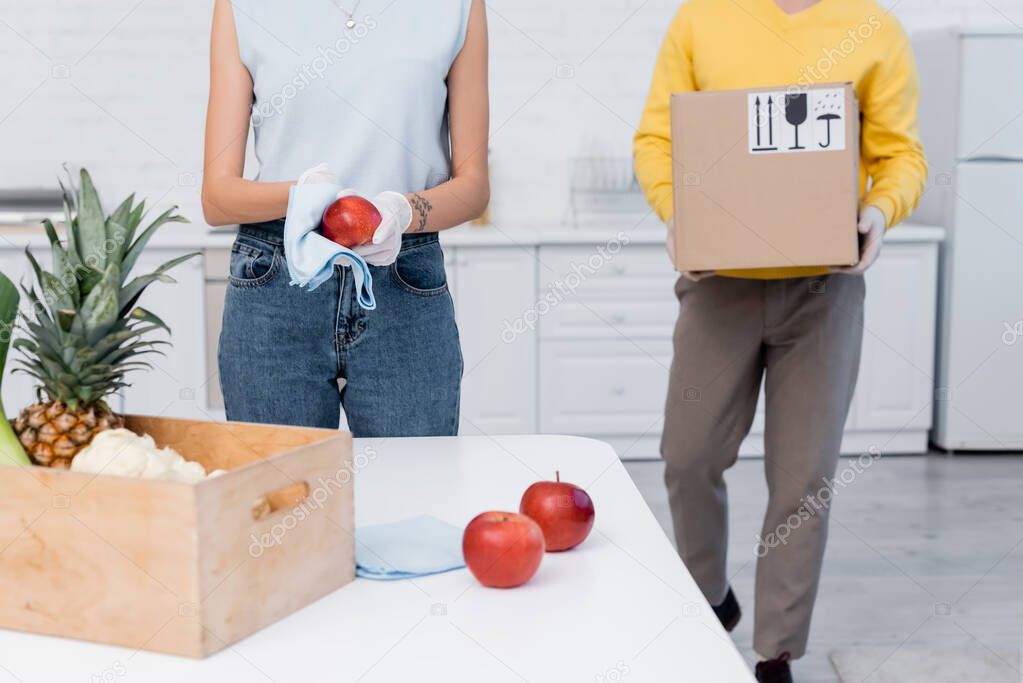 Cropped view of woman in latex gloves cleaning apple near boyfriend carrying carton box in kitchen 