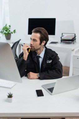 Siyah üniformalı ciddi bir güvenlik görevlisi ofisteki cihazların yanında telsizle konuşuyor.