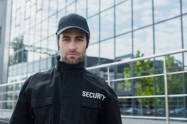 Siyah üniformalı genç güvenlik görevlisi cam cepheli binanın yakınındaki kameraya bakıyor.