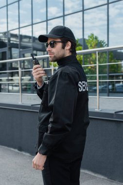 Üniformalı, şapkalı ve güneş gözlüklü genç güvenlik görevlisi binanın yakınında telsizle konuşuyor.