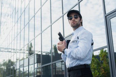 Güneş gözlüklü güvenlik elemanı cam cepheli bir binanın yanında duruyor.