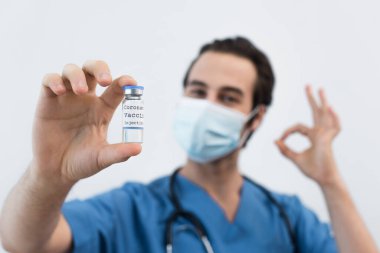 Koronavirüs aşısı taşıyan bulanık maskeli bir doktor ve gri renkte izole edilmiş bir jest gösteriyor.