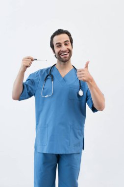 Elektronik termometresi olan memnun doktor gri üzerinde baş parmağını gösteriyor.