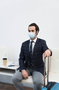 Tıbbi maskeli bir adam kalkış salonunda oturuyor seyahat çantası ve kahvenin yanında.