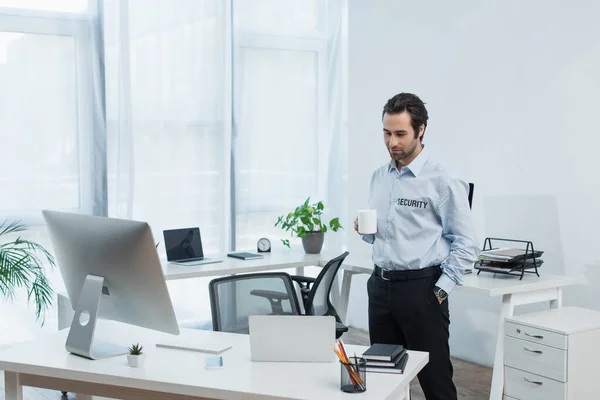 smiling security man with tea cup standing with hand in pocket near laptop and computer monitor