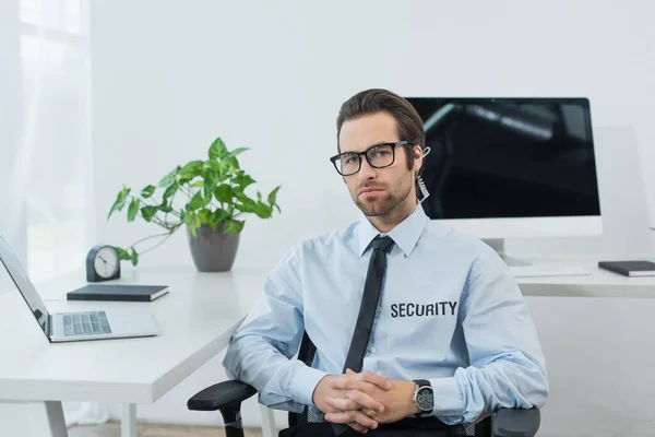 young security man in uniform and earphone looking at camera near computers in supervision room