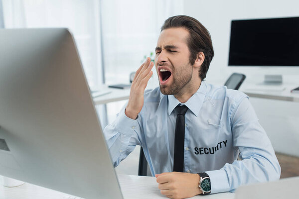 bored security man covering mouth with hand while yawning near blurred computer monitor