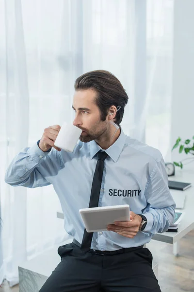 security man in earphone holding laptop and drinking tea in office