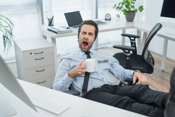 bored guard with cup of tea yawning while sitting with legs on desk in office