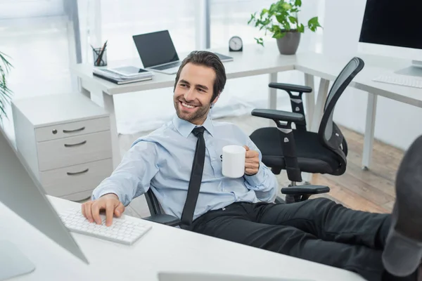 cheerful security man with cup of tea looking at monitor while sitting with legs on desk