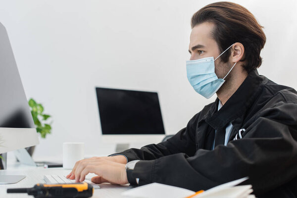 side view of guard in medical mask working on computer near blurred walkie-talkie