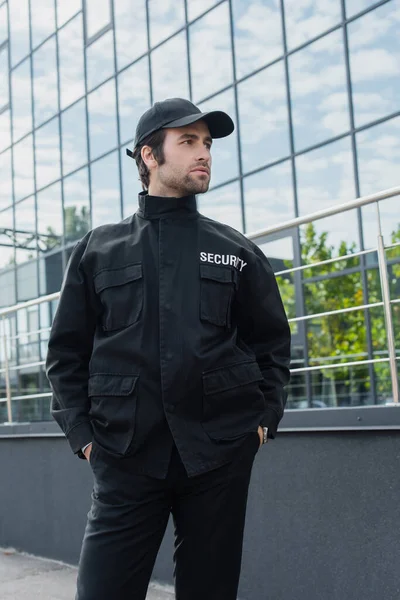 young security man standing with hands in pockets of black uniform and looking away outdoors