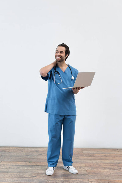 full length of smiling doctor with laptop looking away near grey wall