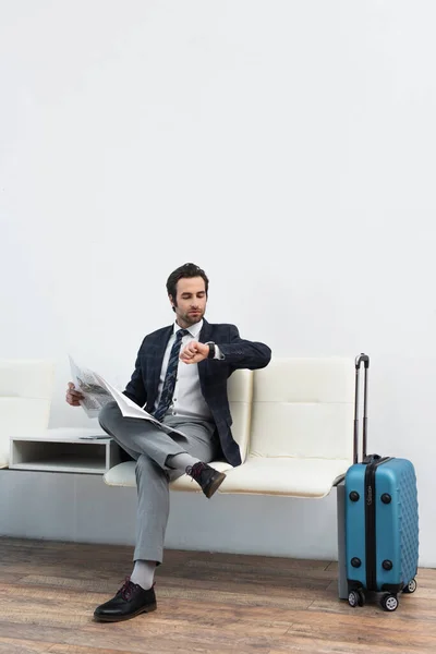 man with newspaper looking at wristwatch while sitting in departure lounge near suitcase