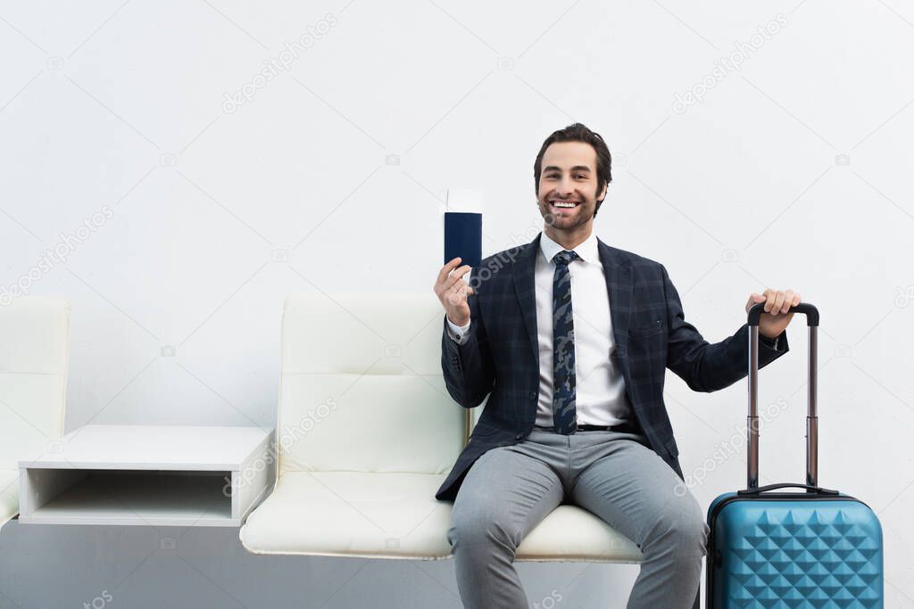 cheerful traveler showing passport and air ticket while sitting in departure lounge near suitcase