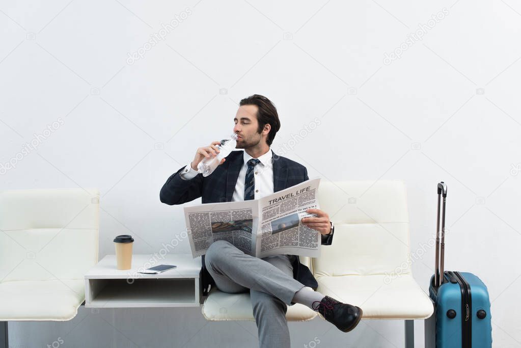 man drinking water while sitting in departure lounge with travel life newspaper