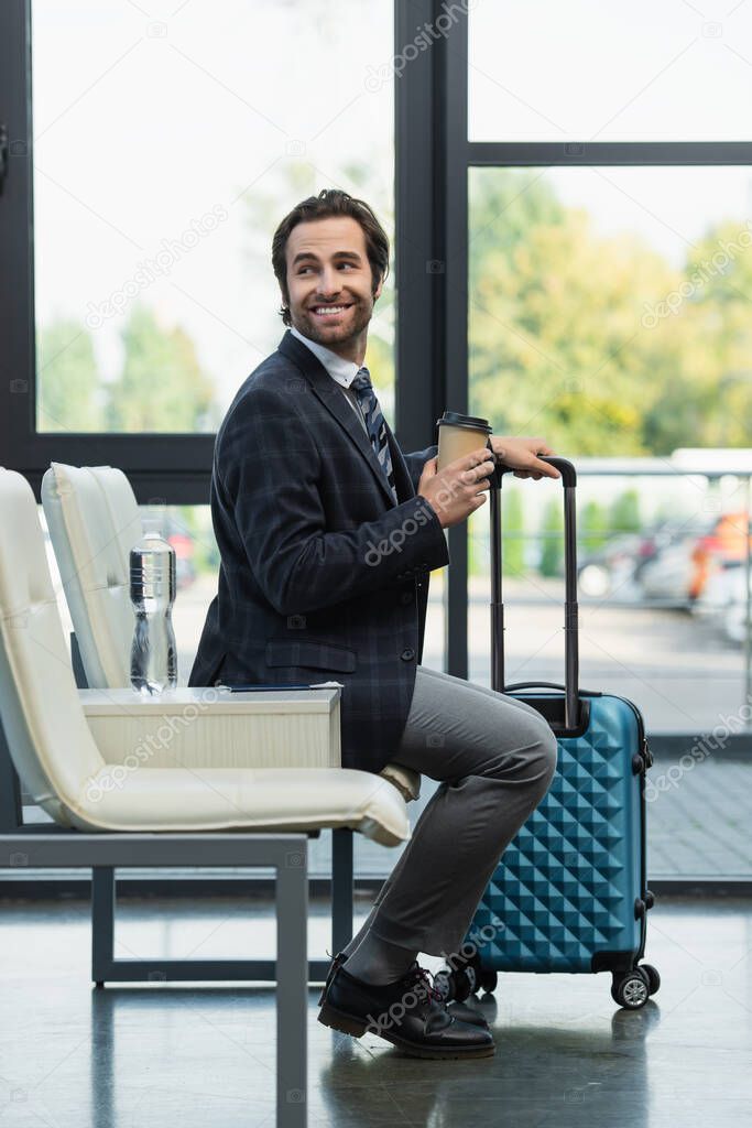 cheerful traveler with paper cup and suitcase sitting in departure lounge and looking away