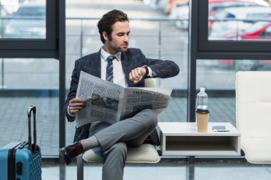 Seyahat hayatı gazetesi olan bir adam kalkış salonunda otururken kol saatine bakıyor.