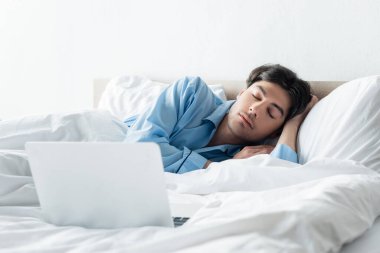 Mavi pijamalı esmer adam sabahları dizüstü bilgisayarın yanında uyuyor.