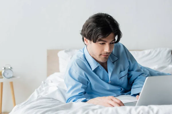 身穿蓝色睡衣的积极男子在笔记本电脑上观看白色被褥上的电影 — 图库照片