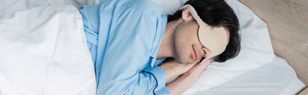 young man sleeping on white bedding in blue pajamas and sleep mask, banner