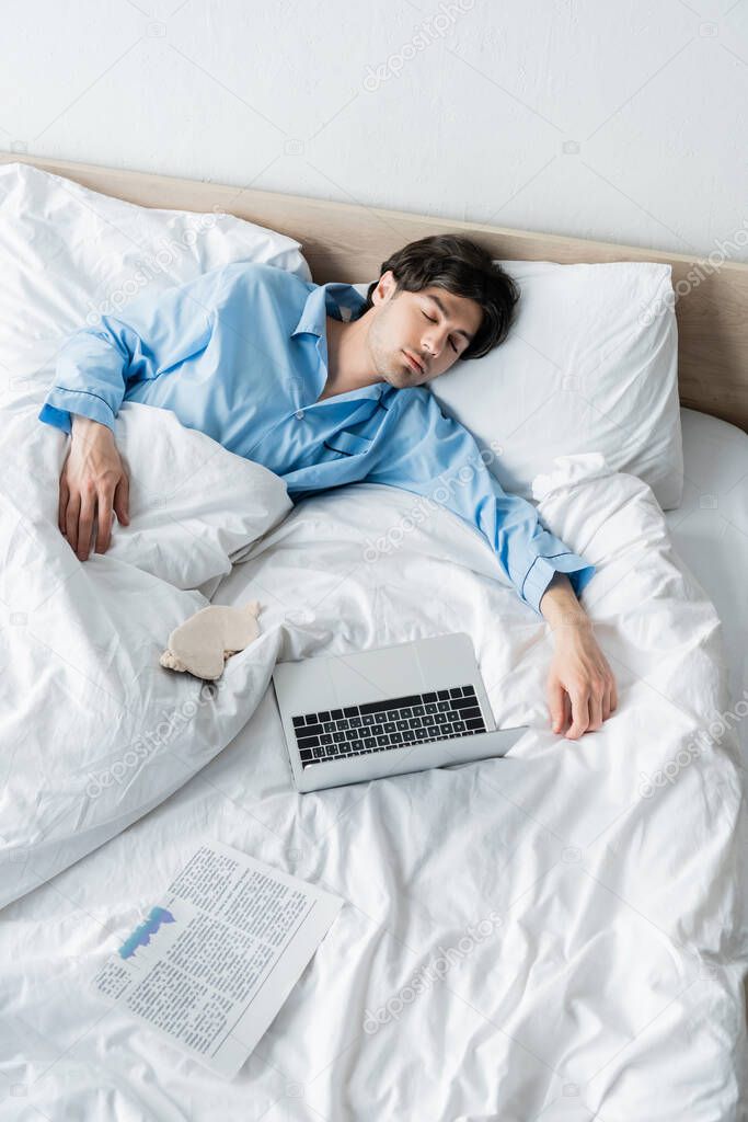 high angle view of man sleeping near laptop, sleep mask and newspaper on bed