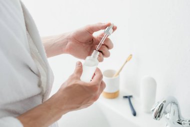 Banyoda kozmetik serumlu şişeyi tutan adamın kırpılmış görüntüsü