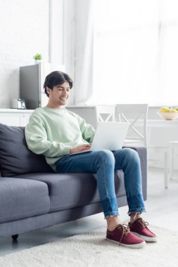 Kanepede dizüstü bilgisayarla otururken gülümseyen esmer adamın tam boy görüntüsü.