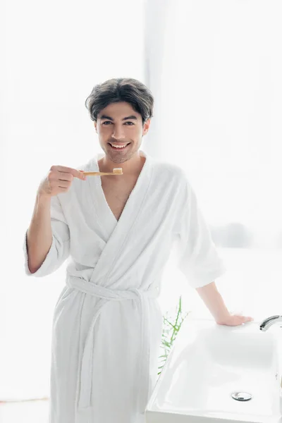 バスルームで歯磨き粉で歯ブラシを持っている間にカメラを見て白いバスローブで楽しい男 — ストック写真