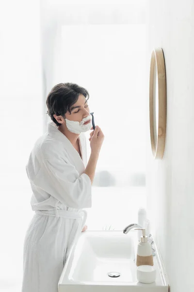 穿着白色浴衣的年轻人在镜子前刮胡子 然后在浴室里洗澡 — 图库照片
