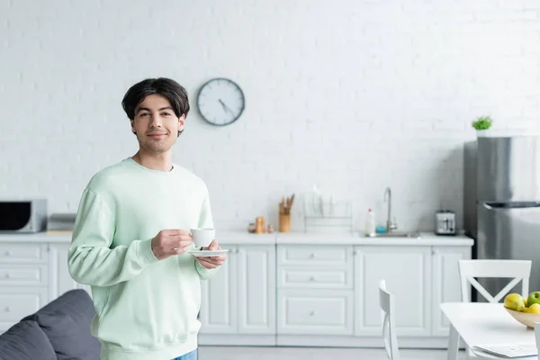 Uśmiechnięta Brunetka Mężczyzna Filiżanką Kawy Patrząc Aparat Fotograficzny Nowoczesnej Kuchni — Zdjęcie stockowe